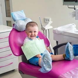 A little boy sitting in a dentist’s chair with coverings over his shoes and a dental bib on waiting for his frenectomy procedure